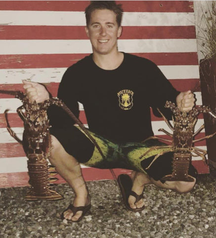 Diver holding two lobsters he harvested. he's kneeling in front of an American Flag wearing a MutinyDiveCo short sleeve black and white t-shirt.