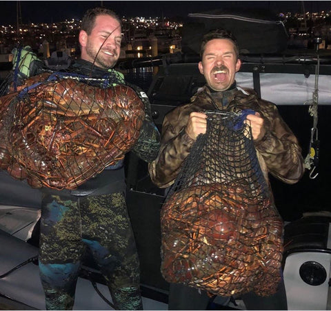Two ecstatic freedivers standing on a dock at night holding massive catches of massive bugs! they are in wetsuits holding their MutinyDiveCo lobster diving bags.